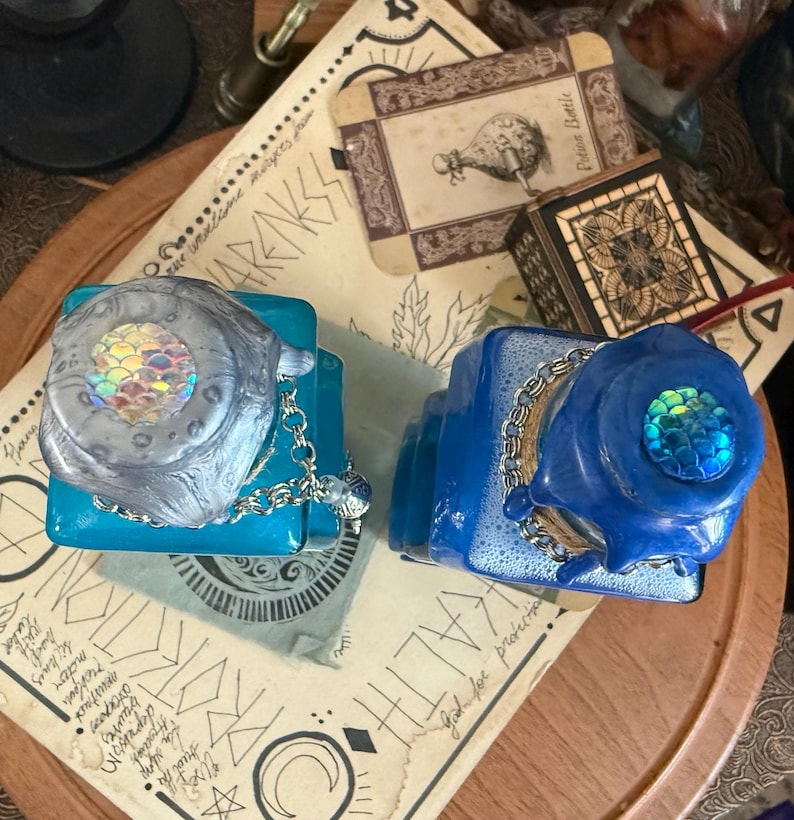 Op de afbeelding: Twee decoratieve glazen flessen met blauwe en turquoise vloeistof. De flessen hebben iriserende flesstoppen met zeemeerminschubben en zilveren kettingen.