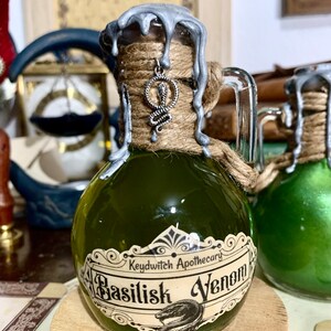 May include: A small glass bottle with a green liquid inside. The bottle is wrapped in twine and has a silver wax seal. The label reads "Keydwitch Apothecary Basilisk Venom".