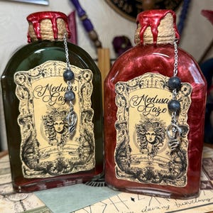 May include: Two glass bottles with cork stoppers and labels that say "Medusa's Gaze" and "Keydwitch Apothecary". The bottles are filled with green and red liquid and have silver chains with black beads hanging from the stoppers. The bottles are sitting on a brown surface with a piece of paper that says "Good for protection during travelling".