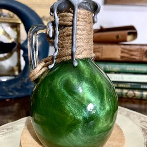 May include: A small glass bottle with a green liquid inside. The bottle has a silver lid with a rope wrapped around it. The lid has drips of silver wax running down the sides.