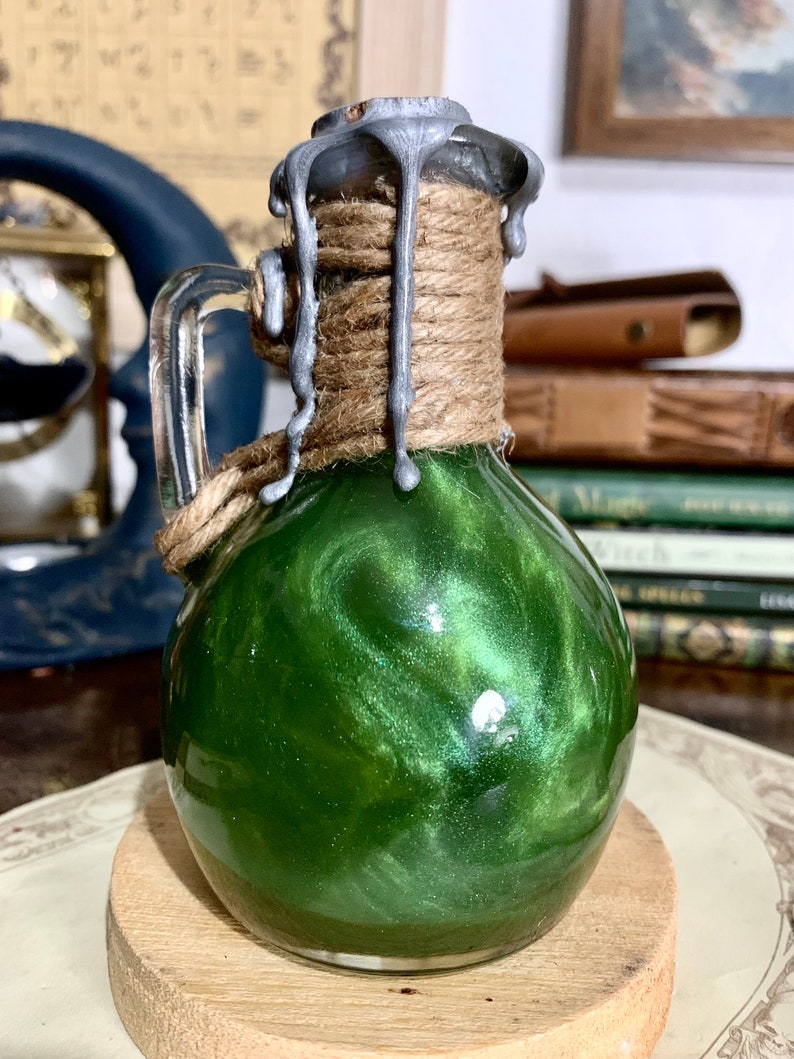 May include: A small glass bottle with a green liquid inside. The bottle has a silver lid with a rope wrapped around it. The lid has drips of silver wax running down the sides.