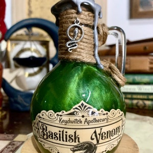 May include: A small green glass bottle with a silver lid and a label that reads "Keydwitch Apothecary Basilisk Venom". The bottle is tied with twine and has a silver snake charm hanging from the lid.