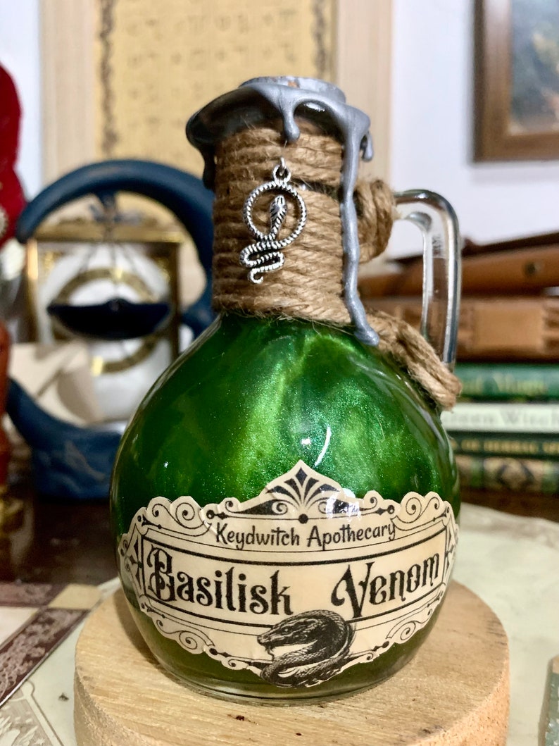 May include: A small green glass bottle with a silver lid and a label that reads "Keydwitch Apothecary Basilisk Venom". The bottle is tied with twine and has a silver snake charm hanging from the lid.