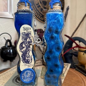 Op de afbeelding: Twee blauwe glazen flessen met een golvend ontwerp, gevuld met een blauwe vloeistof en zwarte glitter. De flessen hebben een blauwe waszegel en een zilveren ketting met een bedel. Het etiket op een flesje staat "Siren Tears" en "Conjuration Alchemy".