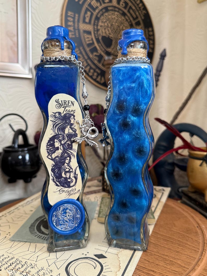Op de afbeelding: Twee blauwe glazen flessen met een golvend ontwerp, gevuld met een blauwe vloeistof en zwarte glitter. De flessen hebben een blauwe waszegel en een zilveren ketting met een bedel. Het etiket op een flesje staat "Siren Tears" en "Conjuration Alchemy".