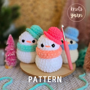 May include: Crocheted snowmen with colorful hats and scarves. The snowmen are white with black eyes and orange noses. One snowman has a pink hat and orange scarf, another has a teal hat and scarf, and the third has a blue hat and scarf. The word "PATTERN" is at the bottom.