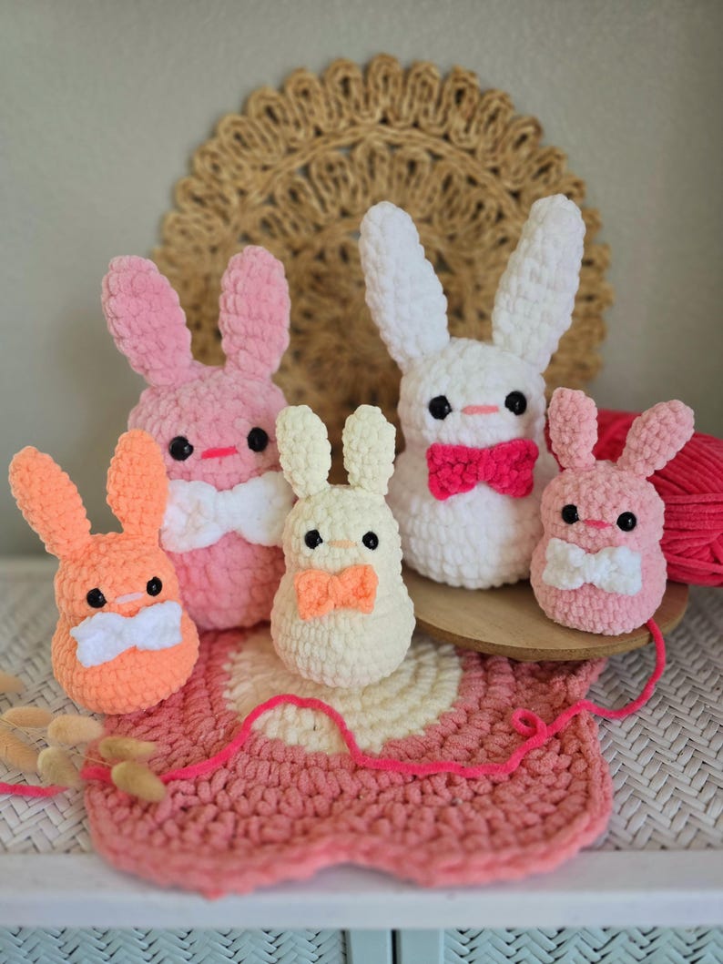 May include: Five crocheted bunny plushies in various colors, including pink, white, and orange. Each bunny has a small bow tie and is sitting on a pink and white crocheted rug.