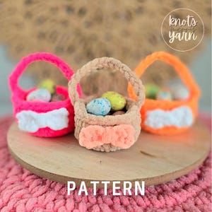 Può includere: Tre colorati cestini pasquali all'uncinetto pieni di uova decorative. I cestini sono rosa, beige e arancioni, ognuno con un dettaglio a fiocco. La parola "PATTERN" è visualizzata nella parte inferiore dell'immagine. Il testo "knots merry yarn" si trova nell'angolo in alto a destra.