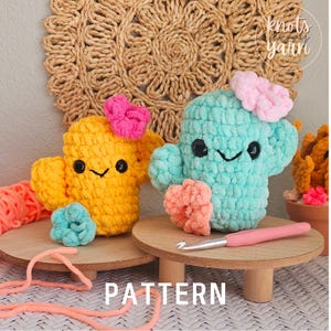 May include: Two crocheted cactus toys, one yellow and one blue, with pink and blue flower accents. The toys are sitting on wooden stands with the text "PATTERN" in front of them.