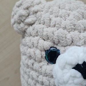 May include: Close-up of a crocheted stuffed animal with a white face and a single blue eye with iridescent glitter. The animal is made with a beige yarn.