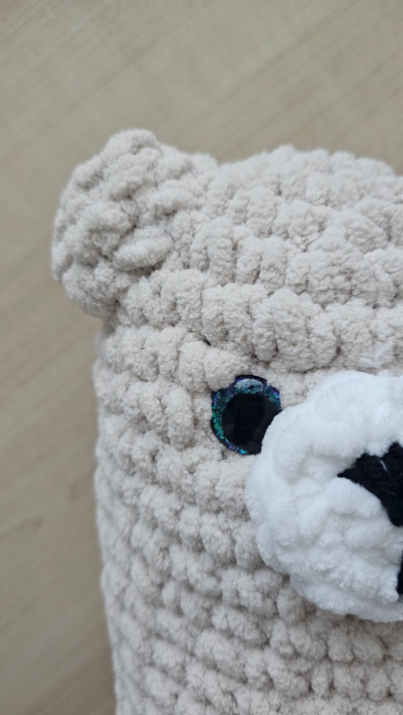 May include: Close-up of a crocheted stuffed animal with a white face and a single blue eye with iridescent glitter. The animal is made with a beige yarn.