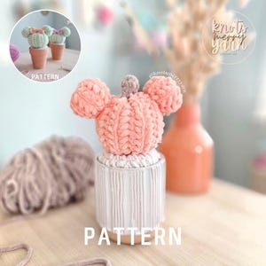 May include: A crocheted peach-colored pumpkin with two rounded ears on top, resembling a mouse shape, sitting in a white ribbed ceramic pot. The pumpkin has a small brown stem and is textured with thick yarn loops. The word PATTERN is visible.