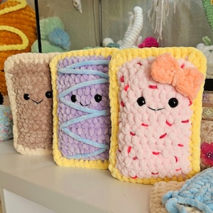 Puede incluir: Tres juguetes de peluche tejidos a crochet con forma de Pop-Tarts con rellenos de diferentes colores y caras sonrientes. Los juguetes est&aacute;n hechos con un hilo suave y peludo y tienen bordes amarillos.