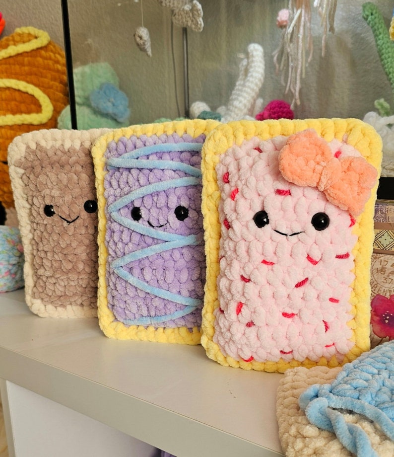Puede incluir: Tres juguetes de peluche tejidos a crochet con forma de Pop-Tarts con rellenos de diferentes colores y caras sonrientes. Los juguetes est&aacute;n hechos con un hilo suave y peludo y tienen bordes amarillos.