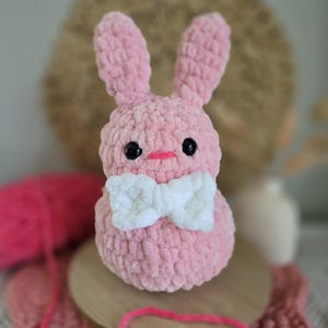 May include: A pink crocheted bunny with a white bow. The bunny has black eyes and a pink nose.