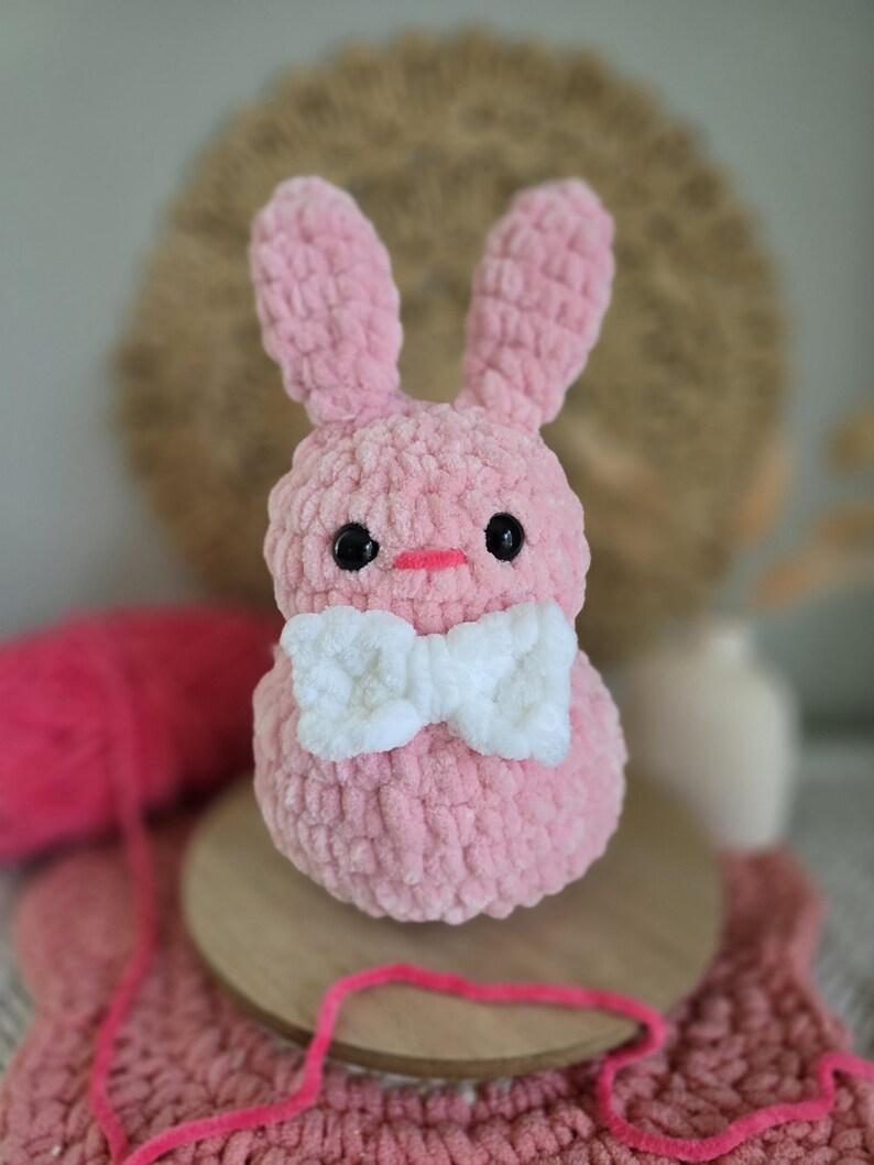 May include: A pink crocheted bunny with a white bow. The bunny has black eyes and a pink nose.
