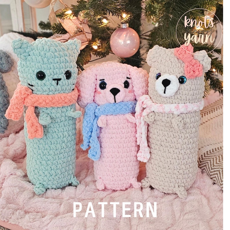 May include: Three crocheted stuffed animals: a light blue cat, a pink dog, and a beige bear. Each animal is wearing a crocheted scarf. The animals are sitting on a pink fuzzy surface. The text "PATTERN" is printed below the animals.