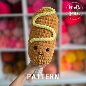 Peut inclure: Un jouet en peluche corndog crocheté avec un motif en spirale jaune et un visage souriant. Le jouet est tenu par une main devant un fond de fil coloré.
