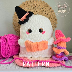 May include: A handmade white crochet ghost wearing a black witch hat with orange trim, holding an orange book. The ghost has large, glittery eyes and pink cheeks. A smaller, stacked witch hat and pink yarn are also present. The word "PATTERN" is visible.