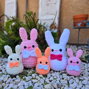 May include: Five crocheted bunny plushies in different colors and sizes. The largest bunny is white with a pink bow, the second largest is pink with a white bow, the third largest is orange with a white bow, and the two smallest bunnies are white and pink with orange bows.