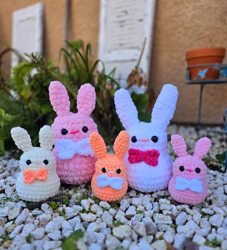 May include: Five crocheted bunny plushies in different colors and sizes. The largest bunny is white with a pink bow, the second largest is pink with a white bow, the third largest is orange with a white bow, and the two smallest bunnies are white and pink with orange bows.