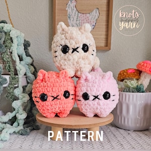 May include: Three crocheted cat plushies in pink, peach, and cream colors. The plushies have black eyes and a stitched-on mouth. The plushies are sitting on a wooden stand with the word "PATTERN" written on it. The background includes a green plant, a wooden sign with the text "knots merry yarn", and a small potted plant.