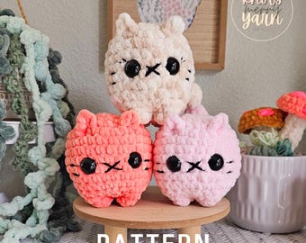 May include: Three crocheted cat plushies in pink, peach, and cream colors. The plushies have black eyes and a stitched-on mouth. The plushies are sitting on a wooden stand with the word "PATTERN" written on it. The background includes a green plant, a wooden sign with the text "knots merry yarn", and a small potted plant.