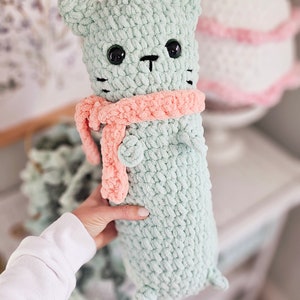 May include: A crocheted green cat plush toy with a pink scarf. The cat has black eyes and a black nose.