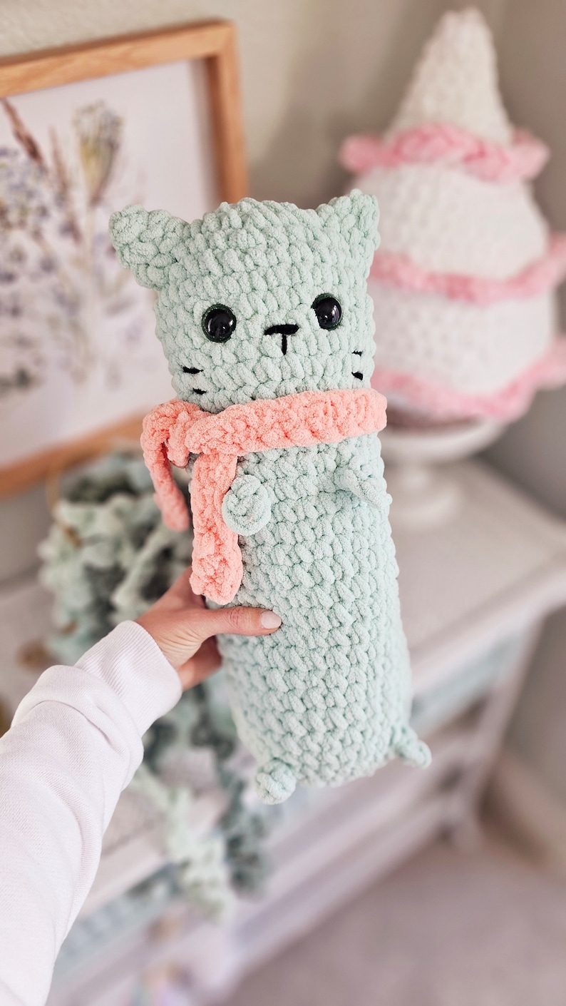 May include: A crocheted green cat plush toy with a pink scarf. The cat has black eyes and a black nose.