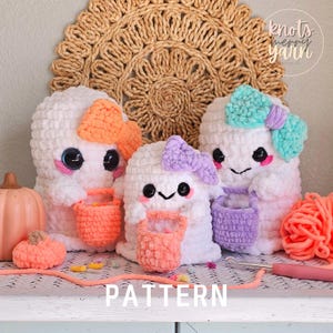 May include: Three crocheted ghost amigurumi toys holding orange, purple, and pink trick-or-treat bags. The ghosts have black eyes, pink cheeks, and are wearing colorful bows. The word "PATTERN" is written in white on a wooden surface below the ghosts.
