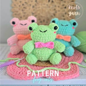 May include: Three crocheted frog toys in pink, green, and teal, each with a bow tie. The green frog is centered, with a pink bow tie. The text "PATTERN frog only" is at the bottom. The toys are on a pink crocheted surface.