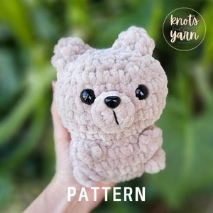 May include: A crocheted plush bear toy in a light brown color with black eyes and a black nose. The bear is being held in a hand with a green background.
