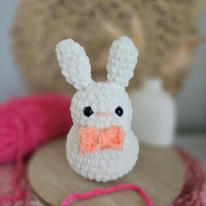 May include: White crocheted bunny with a pink bow tie. The bunny is sitting on a wooden base with pink yarn.