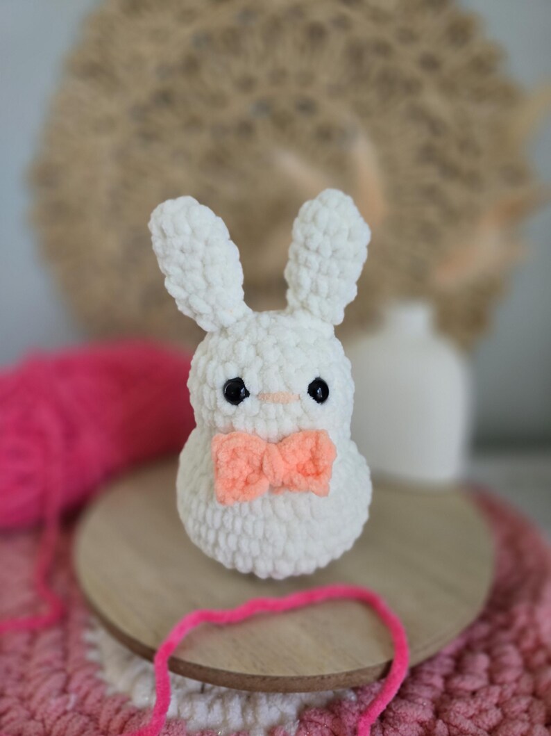 May include: White crocheted bunny with a pink bow tie. The bunny is sitting on a wooden base with pink yarn.
