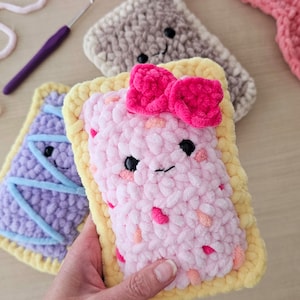 Puede incluir: Una almohada de crochet rosa con forma de pop tart con una corteza amarilla y un lazo rosa en la parte superior. La almohada tiene una cara sonriente con ojos negros y una boca rosa.
