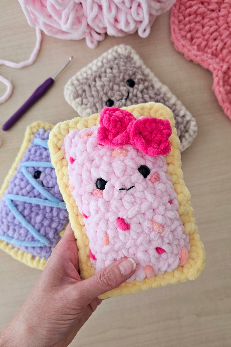 Puede incluir: Una almohada de crochet rosa con forma de pop tart con una corteza amarilla y un lazo rosa en la parte superior. La almohada tiene una cara sonriente con ojos negros y una boca rosa.