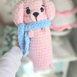 May include: A pink crocheted dog toy with a blue scarf. The toy has black eyes and a black nose.