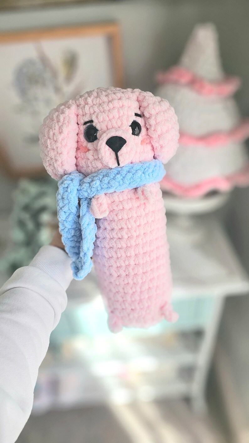 May include: A pink crocheted dog toy with a blue scarf. The toy has black eyes and a black nose.