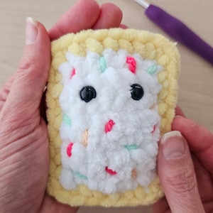 Puede incluir: Un juguete de peluche tejido a crochet con forma de Pop-Tart con un relleno de glaseado blanco y coloridos espolvoreados. El juguete tiene dos ojos negros de bot&oacute;n.