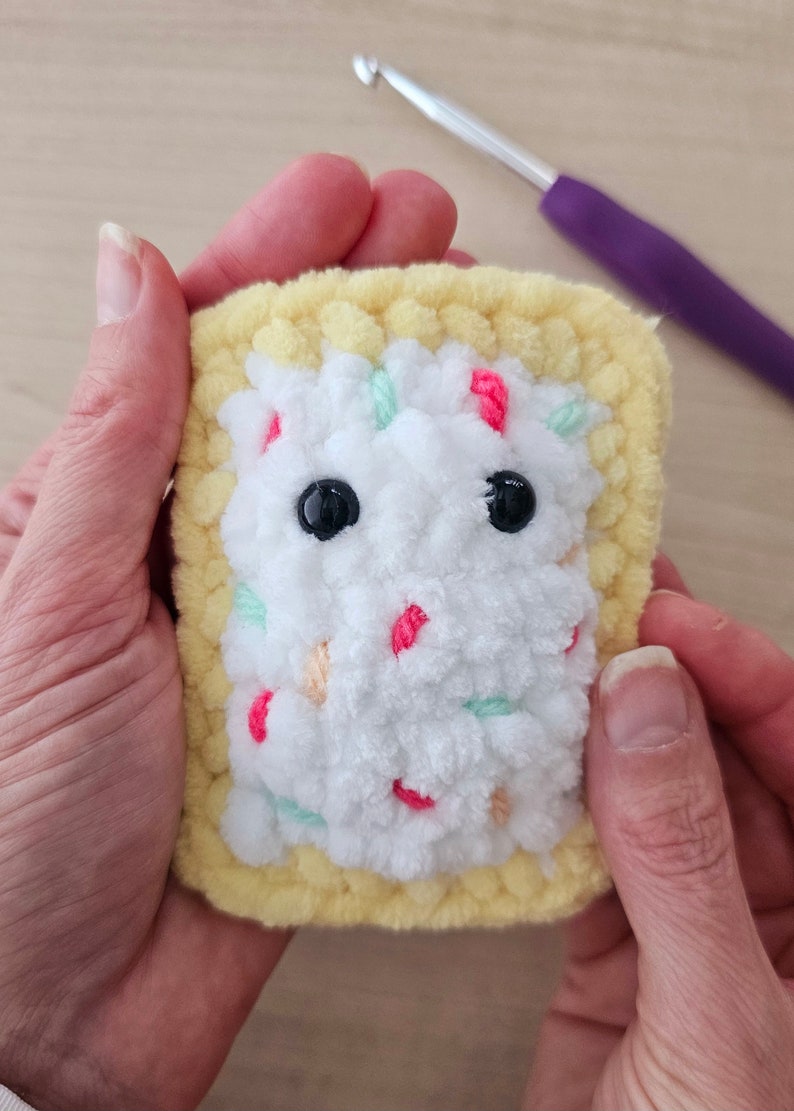 Puede incluir: Un juguete de peluche tejido a crochet con forma de Pop-Tart con un relleno de glaseado blanco y coloridos espolvoreados. El juguete tiene dos ojos negros de bot&oacute;n.