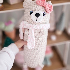 May include: A crocheted plush toy bear with a pink bow and a pink and white scarf. The bear is light beige and has black eyes and a black nose.