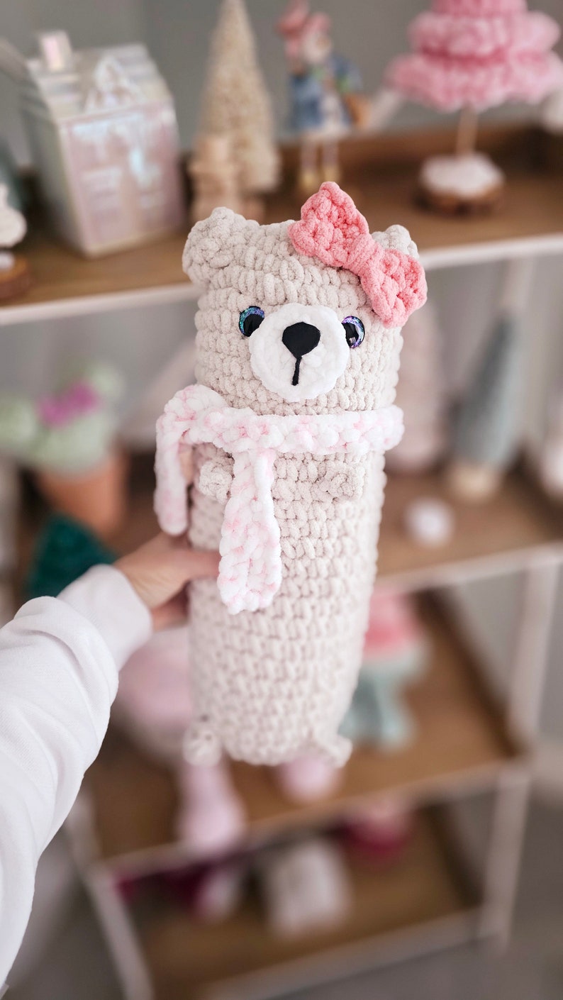 May include: A crocheted plush toy bear with a pink bow and a pink and white scarf. The bear is light beige and has black eyes and a black nose.