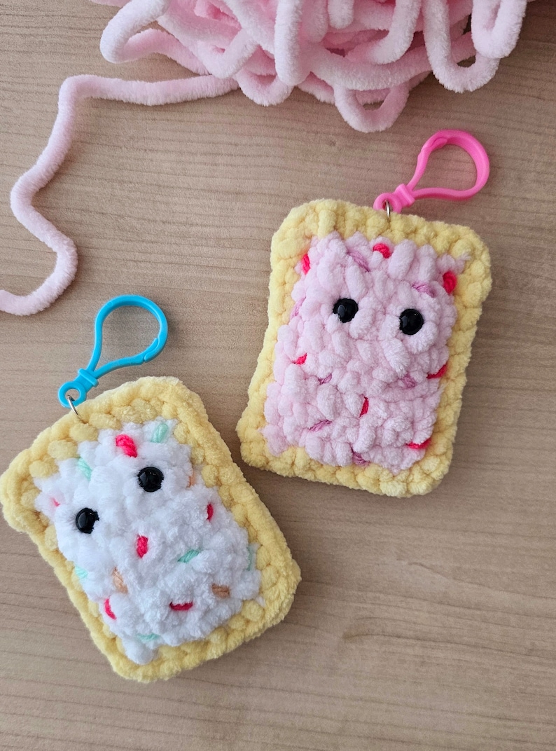 Puede incluir: Dos llaveros de ganchillo con forma de Pop-Tarts. Uno es rosa con chispas rojas y el otro es blanco con chispas rosas y verdes.