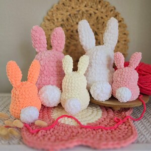 May include: Five crocheted bunny rabbits in different colors, including pink, orange, white, and yellow. The bunnies are sitting on a pink and white crocheted mat.