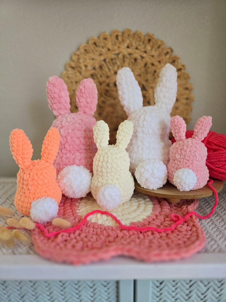 May include: Five crocheted bunny rabbits in different colors, including pink, orange, white, and yellow. The bunnies are sitting on a pink and white crocheted mat.