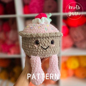 Könnte beinhalten: Ein gehäkeltes Cupcake-Amigurumi-Spielzeug mit rosa Zuckerguss, Streuseln und einer roten Kirsche oben drauf. Der Cupcake hat braune Beine und ein lächelndes Gesicht. Das Spielzeug sitzt auf einer Hand und der Text "PATTERN" ist unter dem Spielzeug sichtbar.