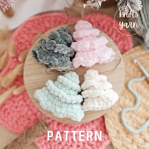 May include: Four crocheted Christmas tree decorations in different colours: grey, pink, light blue, and white. The decorations are arranged on a wooden surface with the text "PATTERN" below them.