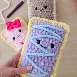Puede incluir: Tres juguetes de peluche tejidos a crochet con forma de tartas pop. Las tartas son de color rosa, morado y marr&oacute;n con glaseado amarillo. La tarta morada tiene glaseado azul en la parte superior.