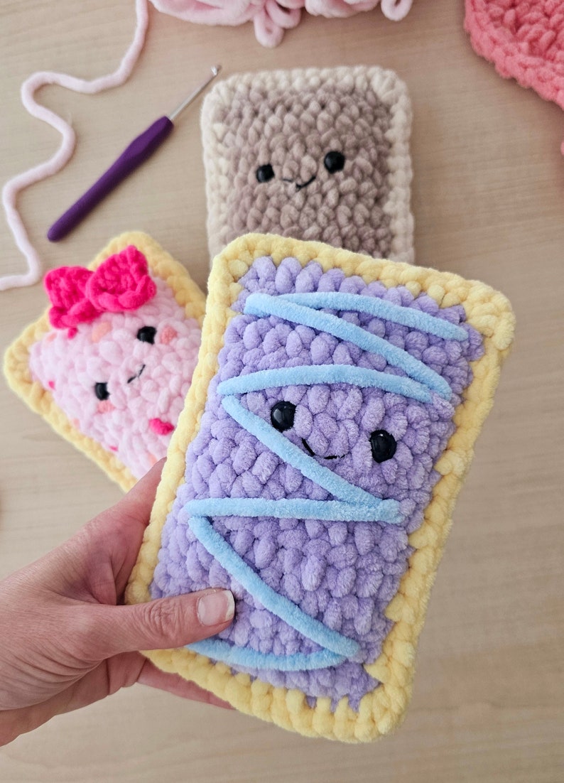 Puede incluir: Tres juguetes de peluche tejidos a crochet con forma de tartas pop. Las tartas son de color rosa, morado y marr&oacute;n con glaseado amarillo. La tarta morada tiene glaseado azul en la parte superior.