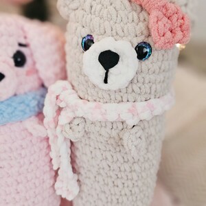 May include: A crocheted gray bear with a pink bow and a pink and white scarf. The bear is sitting on a pink fuzzy surface.
